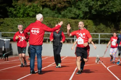 Es sind Läufer auf einer Tartanbahn zu sehen, sie tragen rote Shirts mit Startnummern drauf.