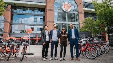 Anjes Tjarks, Esin Rager, Cornelius Kiermasch und Wilken Engelbracht stehen nebeneinander auf dem Vorplatz der Südtribüne jeweils des Millerntor-Stadion, im Hintergrund ist das Millerntor-Stadion zu sehen. Links und rechts von ihnen stehen mehrere Fahrräder.