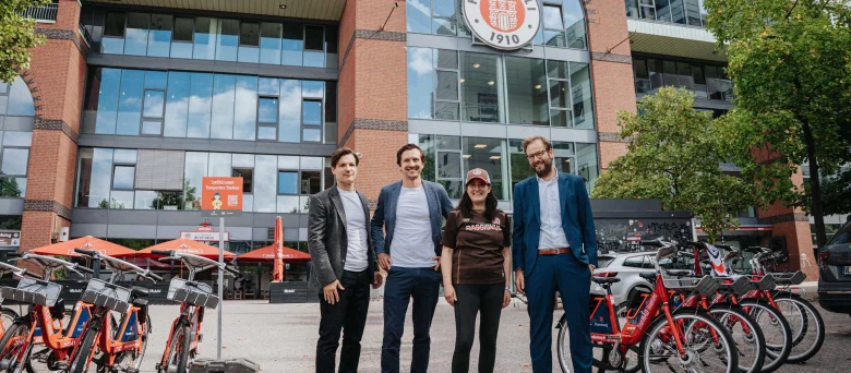 Anjes Tjarks, Esin Rager, Cornelius Kiermasch und Wilken Engelbracht stehen nebeneinander auf dem Vorplatz der Südtribüne jeweils des Millerntor-Stadion, im Hintergrund ist das Millerntor-Stadion zu sehen. Links und rechts von ihnen stehen mehrere Fahrräder.