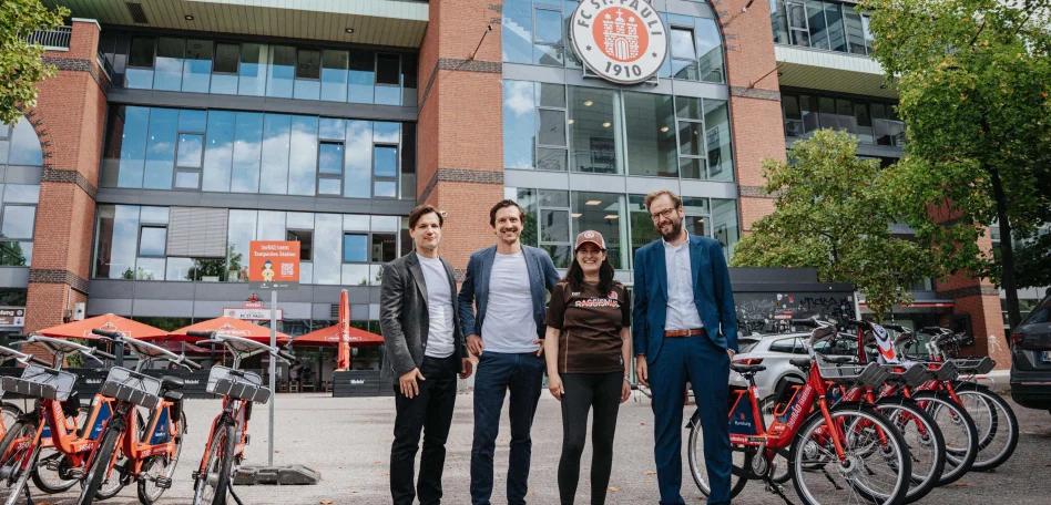 Anjes Tjarks, Esin Rager, Cornelius Kiermasch und Wilken Engelbracht stehen nebeneinander auf dem Vorplatz der Südtribüne jeweils des Millerntor-Stadion, im Hintergrund ist das Millerntor-Stadion zu sehen. Links und rechts von ihnen stehen mehrere Fahrräder.