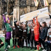 Spieler:innen vom Blindenfußball halten jubelnd einen Pokal in die Luft. Im Hintergrund der Schriftzug "Deutscher Meister".