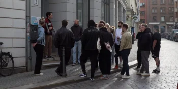 Mehrere Personen sind vor einem Haus im Stadtteil St. Pauli zu sehen, links und rechts sind grafische Elemente in den Farben Schwarz, Rot, Braun und Weiß eingebaut.