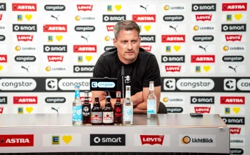 Alexander Blessin sitzt an einem Pult, vor ihm stehen ein Mikrofon und mehrere Flaschen, im Hintergrund ist eine Wand mit verschieden Logos der Sponsoren des FC St. Pauli zu sehen.