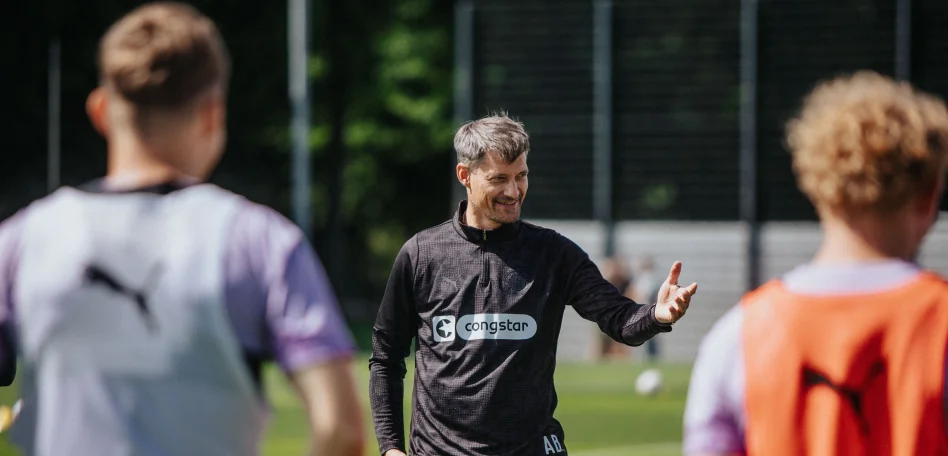 Cheftrainer Alexander hebt die linke Hand und spricht zur Mannschaft, vorne sind von hinten zwei Spieler unscharf zu erkennen.