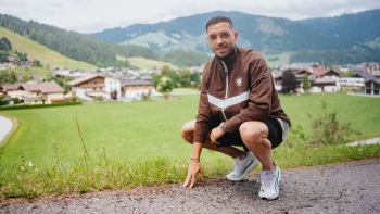 Mathias Pereira Lage hockt auf einem Weg, er trägt eine braun-weiße Trainingsjacke. Im Hintergrund ist Alpenpanorama zu sehen.