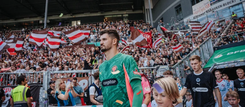 Nikola Vasilj beim Einlaufen ins Millerntor-Stadion, im Hintergrund sind Fans auf der Tribüne und weitere Spieler zu sehen.