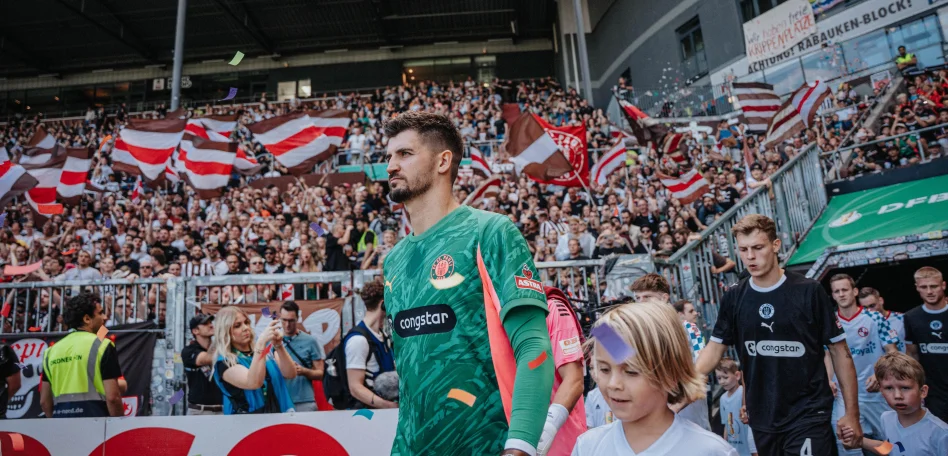 Nikola Vasilj beim Einlaufen ins Millerntor-Stadion, im Hintergrund sind Fans auf der Tribüne und weitere Spieler zu sehen.