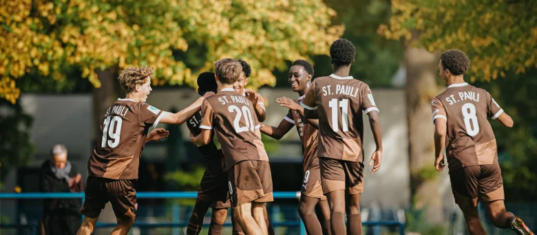 Jubelnde Jugendfußballspieler des FC St. Pauli in braunen Trikots feiern gemeinsam ein Tor auf dem Spielfeld, umgeben von herbstlich gefärbten Bäumen.