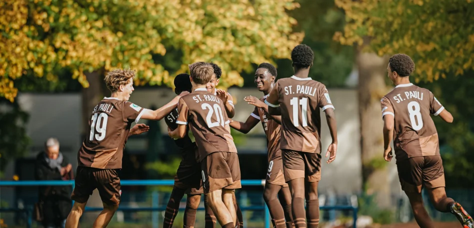 Jubelnde Jugendfußballspieler des FC St. Pauli in braunen Trikots feiern gemeinsam ein Tor auf dem Spielfeld, umgeben von herbstlich gefärbten Bäumen.