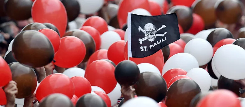 Eine Menschenmenge die Ballons in rot, braun und weiß hochhält. Mittendrin eine kleine St. Pauli Flagge mit Totenkopf
