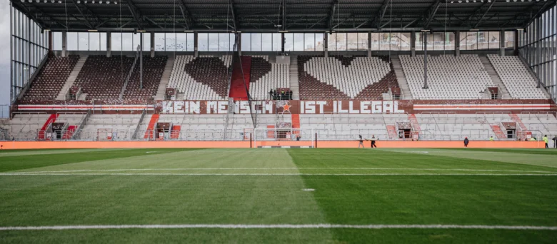 Ein Bild vom Stadion von innen. Die Stühle bilden zwei Herzen und ein Banner zeigt den Spruch "Kein Mensch ist illegal"