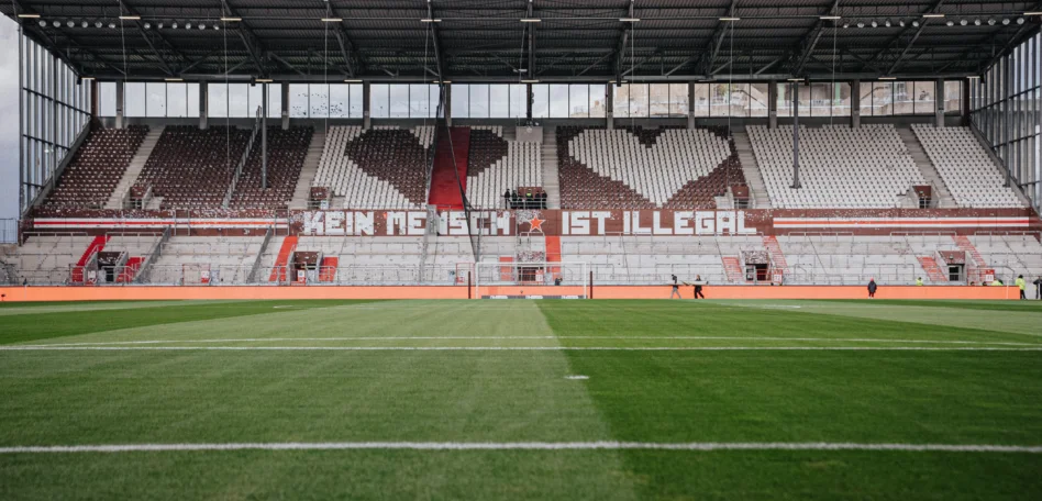 Ein Bild vom Stadion von innen. Die Stühle bilden zwei Herzen und ein Banner zeigt den Spruch "Kein Mensch ist illegal"