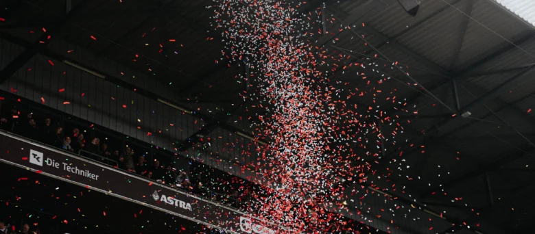 Man sieht eine Tribünenseite eines vollen Millerntor Stadions. Ein rot weißer Konfetti Regen fliegt durch die Luft