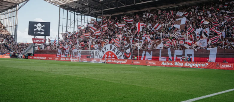 Man sieht eine Seite des vollen Stadions. Fahnen werden geschwungen und Konfetti und Krepppapier liegt auf dem Boden