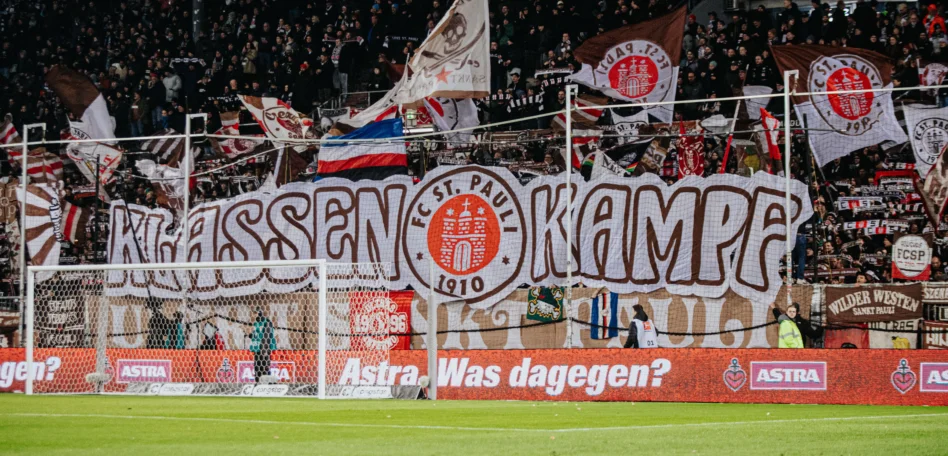 Man sieht die Fankurve mit vielen St Pauli Flaggen und Bannern. Auf einem steht groß geschrieben "Klassenkampf".