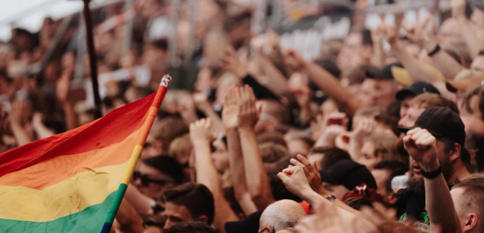 Man sieht einen Ausschnitt des vollen Fanblocks. Eine Regenbogenfahne wird geschwungen. Die Menschen strecken ihre Faust in die Höhe.