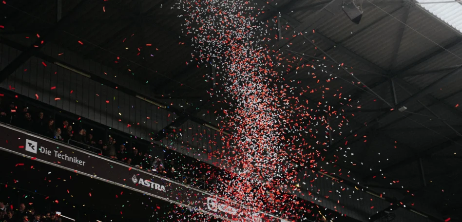 Man sieht eine Tribünenseite eines vollen Millerntor Stadions. Ein rot weißer Konfetti Regen fliegt durch die Luft