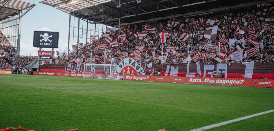 Man sieht eine Seite des vollen Stadions. Fahnen werden geschwungen und Konfetti und Krepppapier liegt auf dem Boden