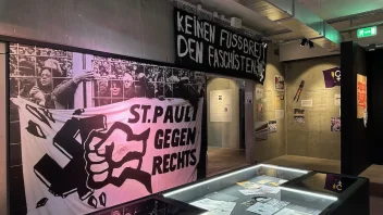 Es ist ein Teil des FC St. Pauli-Museums zu sehen, vorne ein Vitrine, im Hintergrund hängen mehrere Fahnen an der Wand.