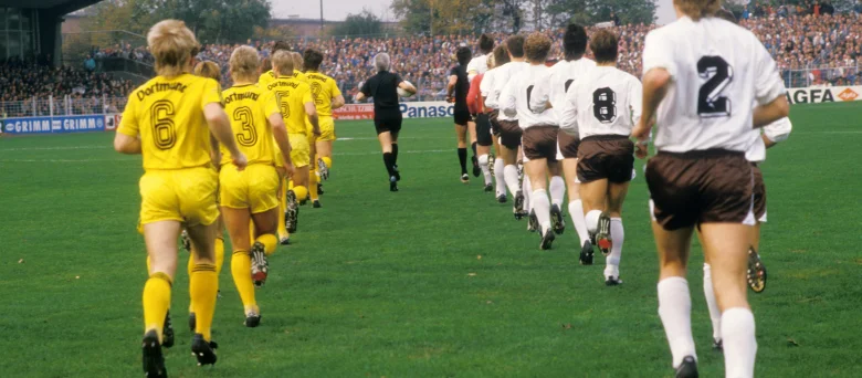 Die Mannschaften des FC St. Pauli und Borussia Dortmund laufen auf den Rasen des Millerntor-Stadions, alle Spieler sind von hinten zu sehen.
