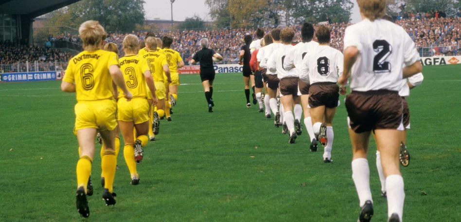 Die Mannschaften des FC St. Pauli und Borussia Dortmund laufen auf den Rasen des Millerntor-Stadions, alle Spieler sind von hinten zu sehen.